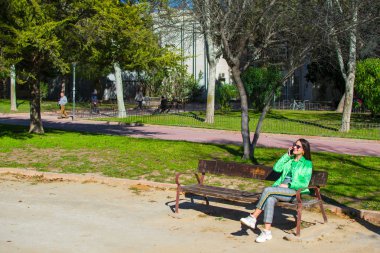 Güzel genç kadın gülümseyerek ve parkta bir bankta otururken cep telefonu kullanarak çekim.