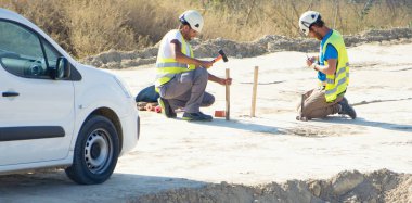 Minibüslerinin yanındaki sopayla zemini ölçen iki inşaat işçisi..