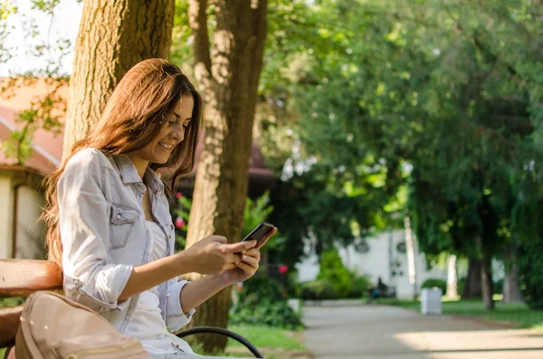 Pretty long hair brunette girl sitting on a bench in a sunlit park and looking at her smart phone.