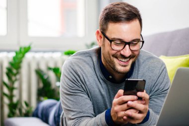 Genç adam gözlüklü telefon ev raslantı kullanırken gülümseyen bir koltukta yalan