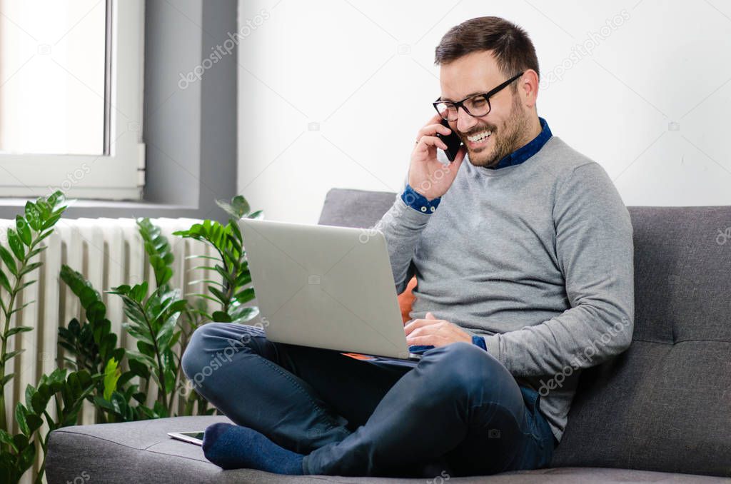 Handsome young man sitting cross-legged on sofa at home, laptop on the knees, talking on the phone and smiling cheerfully. Freelancer