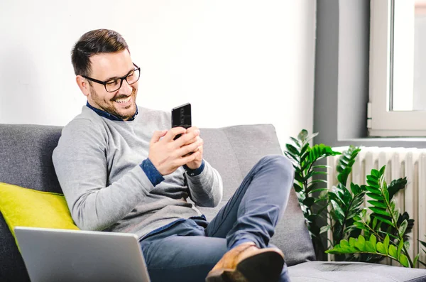 Neşeli smmiling genç adam cep telefonu ve laptop özelliğini kullanarak evinden iletişim. Serbest yaşam tarzı.