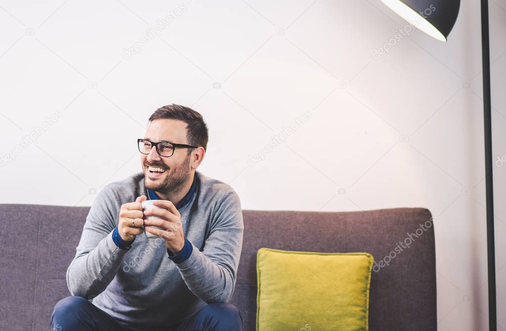 Cheerful handsome young man sitting on sofa, holding hot beverage, smiling and looking away. Leisure time at home. 