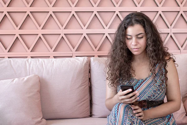 Curly Girl sitter på en rosa soffa surfar hennes telefon — Stockfoto