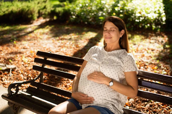 Mujer embarazada disfrutando del descanso en un banco en un parque al atardecer — Foto de Stock