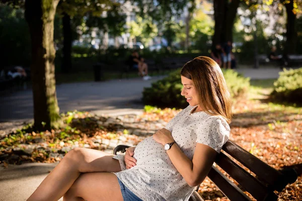 子供を期待している若い女性は、日当たりの良い公園に座っています — ストック写真