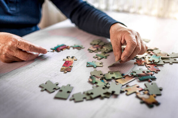 Closeup of the old hands playing with jigsaw puzzles at the white table at home. Connecting pieces together. 