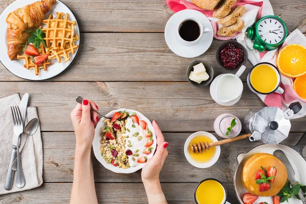 Food and drink, healthy morning eating concept. Breakfast assortment with pancakes, waffles, croissant sandwich and granola on the wooden table. Top view flat lay background with woman hands