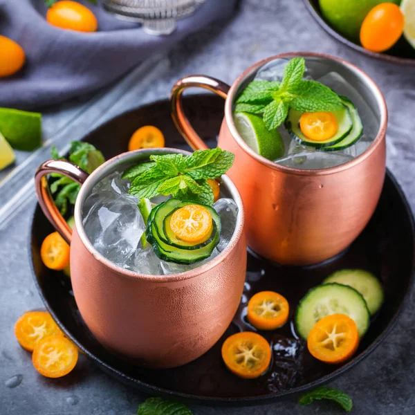 Moscow mule cocktail with green lime, mint, ice, cucumber, kumquat in a copper mug on a black table.