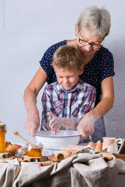 Happy senior mature woman, grandmother and young boy, grandson cooking, kneading dough, baking pie, cake,  cookies. Family time in the cozy kitchen. Autumn activity at home.