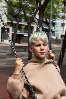 Handsome teenager swinging at the playground outdoors. Adolescent spending the downtime in the city 