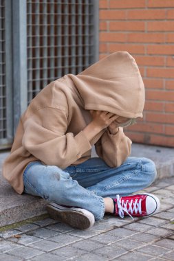 A Sad Lonely teenager hiding his face and crying while sitting alone on the floor outdoors. Young boy struggling with inner problems. Homelessness, Loneliness, bullying, depression and sadness concept
