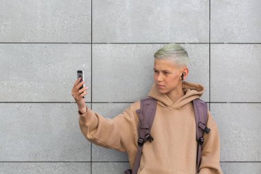 Handsome teenager saying hello standing against grey wall on the city street, outdoors. Caucasian young boy is talking with his friends on the phone