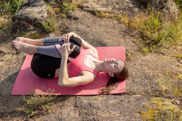 Young smiling caucasian woman lying on the mat,meditating,practicing yoga pose knees to the chest on the rock.Girl wearing pink sportswear,relaxing with namaste.Healthy lifestyle.Copy space