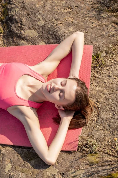 Portrait of young smiling caucasian woman lying on the yoga mat on the rock,taking sunbath,relaxing after meditation,practicing yoga pose.Girl wearing pink sportswear.Healthy lifestyle.Copy space
