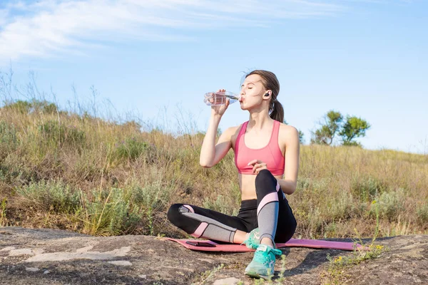 O tânără caucaziană care stă pe covorul de yoga, bea apă din sticlă cu căști mobile Fotografie de stoc