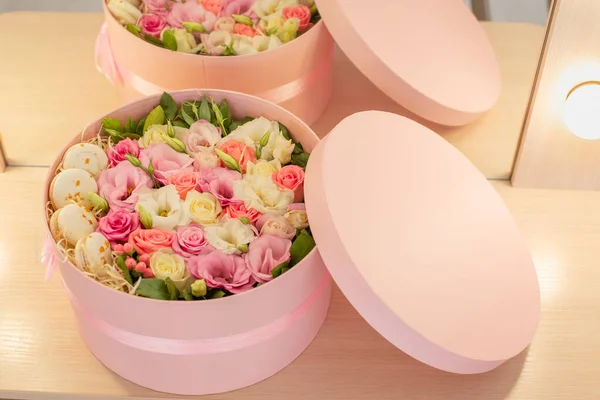 Close up of round pink box with flowers,french macarons and top reflected in the mirror with lights on dressing table.Romantic bouquet,floral composition of rose,eustoma.Florist make wedding decor.