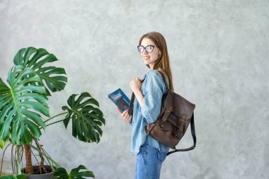 Kahverengi sırt çantalı gülümseyen kız çocuğu ve arka planda canavar bitkisi olan gri duvar arkaplanına izole edilmiş kitaplar. Kot elbise giyen kadın. Üniversite, üniversite, üniversite eğitimi..