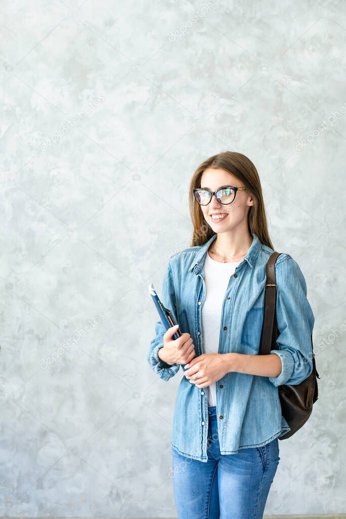 Smiling caucasian girl in glasses holding brown backpack and books isolated on grey wall background indoor.Student wearing denim clothes.Distance education in university,college.Lifestyle.Copy space
