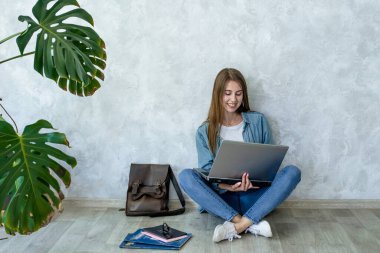 Dizüstü bilgisayar kullanarak gülümseyen genç bir kız, gri arka planda bacak bacak üstüne atarak yerde oturuyor. Kot pantolon giyen bir kadın. Kahverengi sırt çantası, notlar, yakınlarda duran kitaplar. Uzaklık eğitimi..