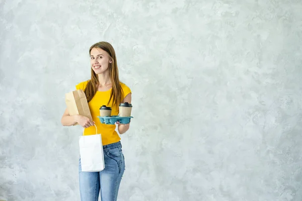 Woman in yellow uniform deliver food isolated on grey background.Smiling courier holding paper bags,cups of coffee.Safe delivery goods from store,restaurant to your home while quarantine.coronavirus