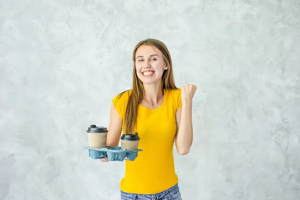 Woman customer receive food delivery,holding two cups of coffe isolated on grey background.Happy girl raised arm,smiling for success.Safe delivery goods from restaurant to your home while quarantine