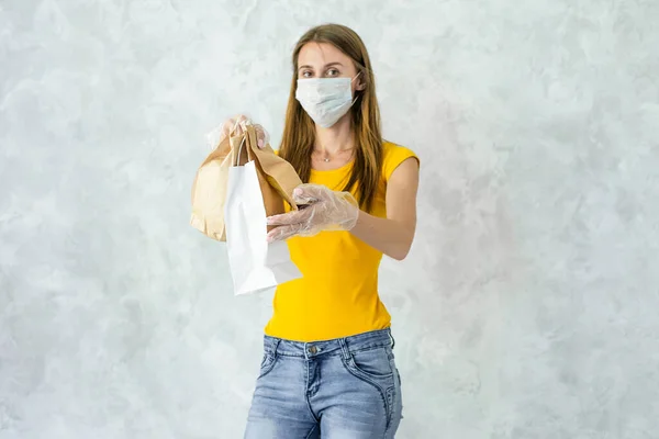 Woman in yellow uniform,protection mask,gloves deliver food isolated on grey background.Courier giving paper bags.Safe delivery goods from store,restaurant to your home while quarantine.coronavirus