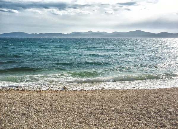 Dalgalar ve çakıl plajı ile arka planda dağlar denize sıfır.