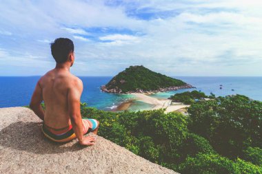 Maço üstsüz bir adam kayanın tepesinde oturmuş deniz manzarasını izliyor, Koh Tao, Tayland