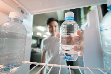 Asyalı bir adam buzdolabında plastik şişeler dolusu içme suyu depoluyor, buzdolabının içini görüyor, geniş açılı bir bakış açısı var.