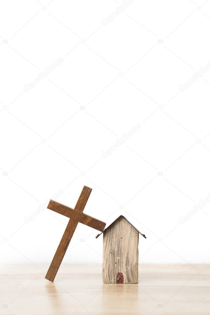 Wooden cross leaning on a small house wooden model on wooden board and white background