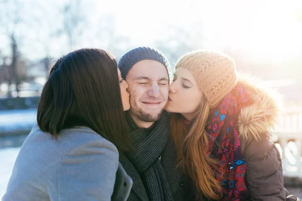 Two beautiful young women kissing handsome man standing between them. Winter outdoors fun