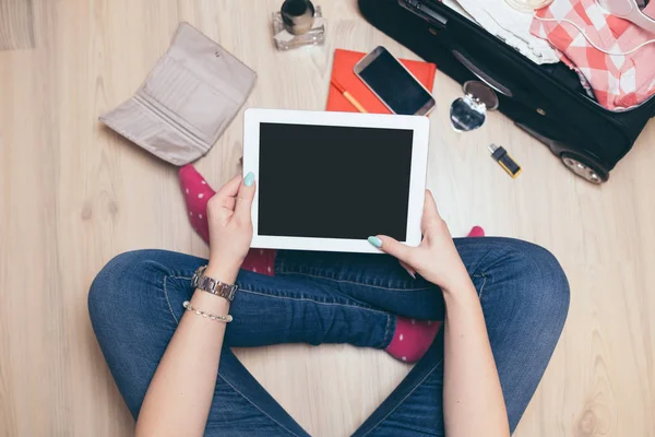 Kadın tableti tutuyor ve seyahat için hazırlanıyor. İçinde kadın kıyafetleri, aksesuarlar, kredi kartları, güneş gözlükleri ve akıllı telefon bulunan gezgin çantası.. 