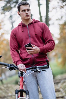 Sonbahar açık havada. Bisiklet Park ile ayakta, cep telefonu tutan kulaklık ile genç ciddi yakışıklı adam. Seçici odak.