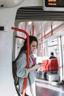 Tramvayda ayakta ve cep telefonunda bir şeyler yapıyor güzel genç kadın.
