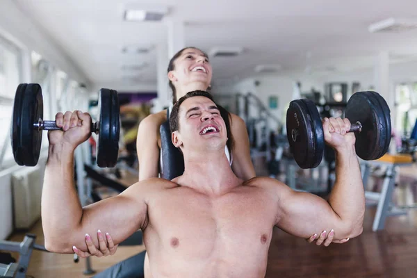 A man and a woman practicing together in the gym and laughing together while woman is helping man in lifting weights.