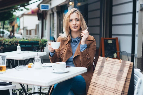 Beautiful blond woman sitting alone in cafeteria, smoking a cigarette and drinking coffee
