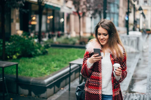 コーヒーの上を歩いて通り 携帯電話のテキスト メッセージのファッショナブルな美しい若い女性 — ストック写真