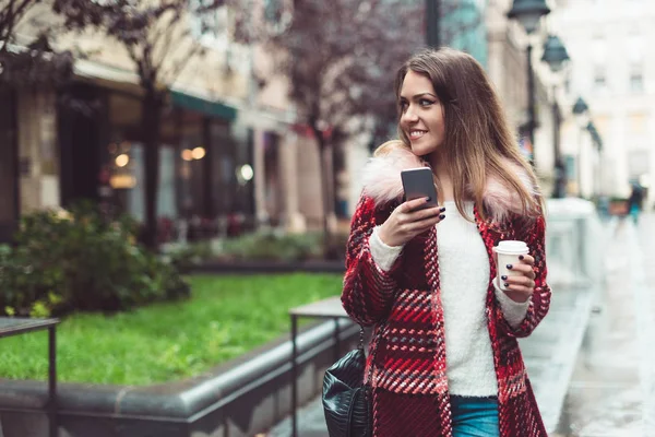 コーヒーの上を歩いて通り 携帯電話のテキスト メッセージのファッショナブルな美しい若い女性 — ストック写真
