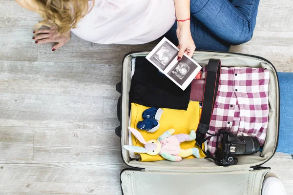Pregnant woman next to open traveler\'s bag with ultrasound scan of her baby, baby socks, clothing and photo camera. Travel and vacations concept.