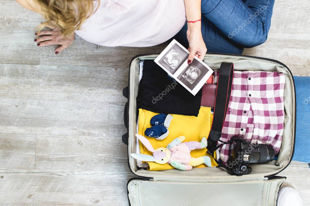 Pregnant woman next to open traveler's bag with ultrasound scan of her baby, baby socks, clothing and photo camera. Travel and vacations concept.