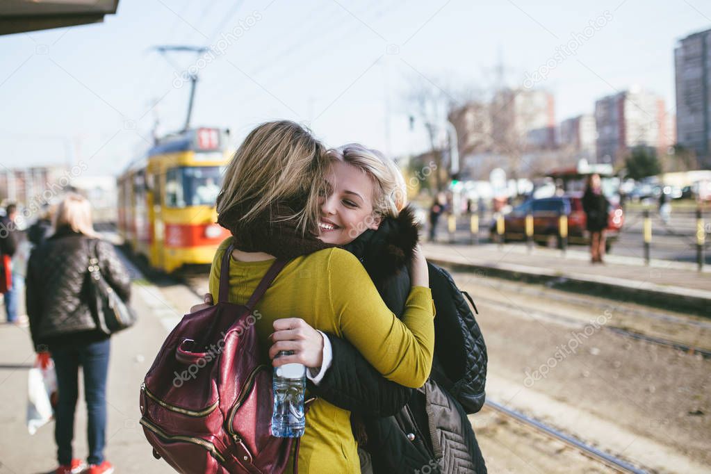 Two beautiful young women friends meet each other after long time at tram station. They smiling and hugging.
