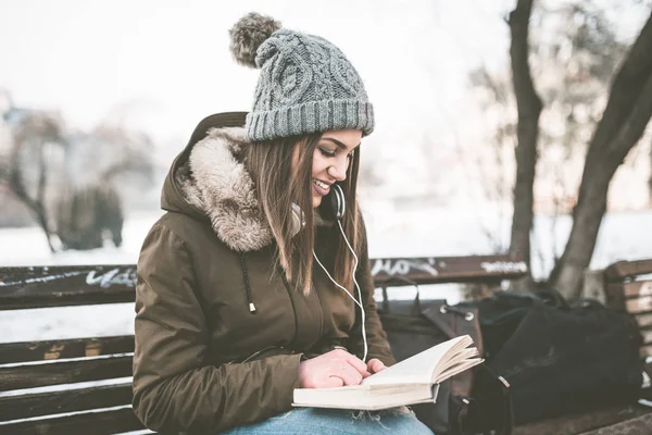 公園のベンチに座って 音楽を聞いて 彼女はノートに何かを書くのヘッドフォンで若くてきれいな女性 — ストック写真