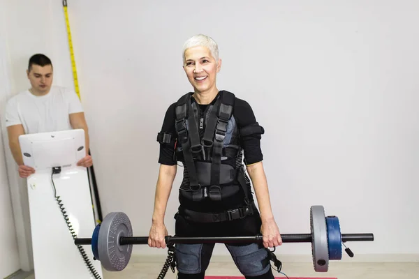 Beautiful senior woman doing exercises in electrical muscular stimulation suit with her personal trainer at rehabilitation center