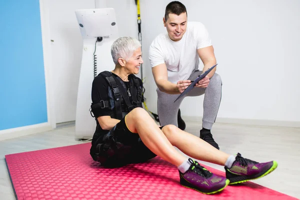 Beautiful senior woman doing exercises in electrical muscular stimulation suit with her personal trainer at rehabilitation center