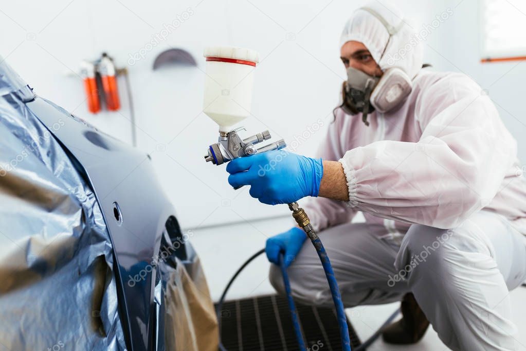 Man with protective clothes and mask painting car using spray compressor. Selective focus.