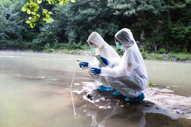 Two scientists in protective suits taking water samples from the river. clipart