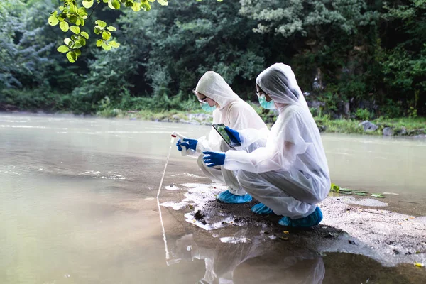 保護スーツを着た2人の科学者が川から水のサンプルを採取 — ストック写真