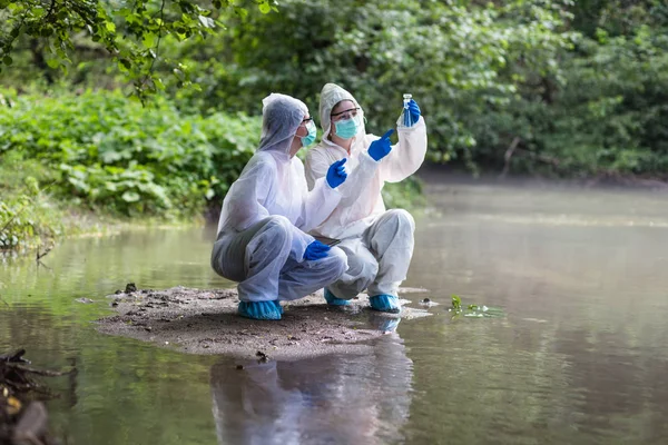 保護スーツを着た2人の科学者が川から水のサンプルを採取 — ストック写真