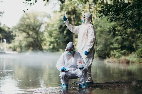 保護スーツを着た2人の科学者が川から水のサンプルを採取 — ストック写真
